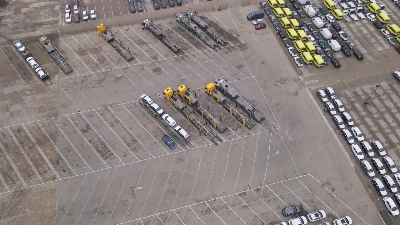 A high-angle video of a parking lot featuring multiple transport trucks with car carriers, parked vehicles, and rows of yellow vans. The drone flies around the vehicles.