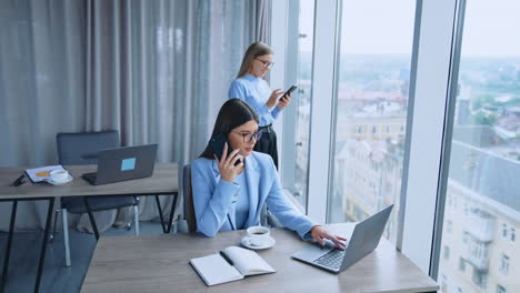 office employees having productive day at work. female colleagues using phones and laptop. office with panoramic windows at backdrop.