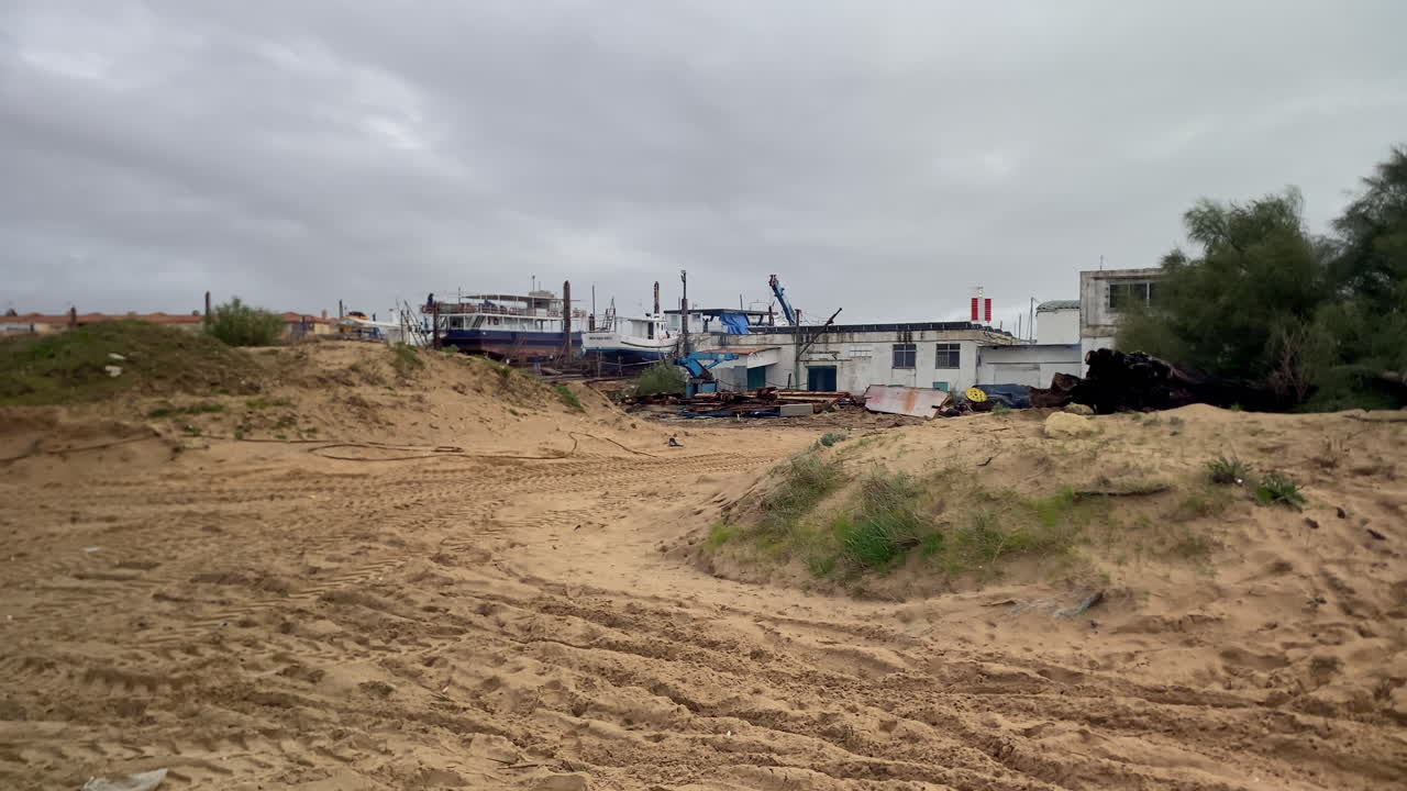 Cantiere navale costiero che espone imbarcazioni in riparazione sulla terraferma, con terreno sabbioso ed edifici industriali sotto un cielo coperto.