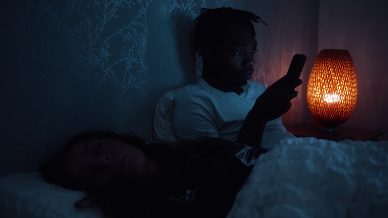 Uomo che usa lo smartphone a tarda notte mentre il partner dorme nel letto accanto a lui