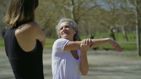 Sporty-senior-and-young-women-stretching-arms-and-talking-in-park.