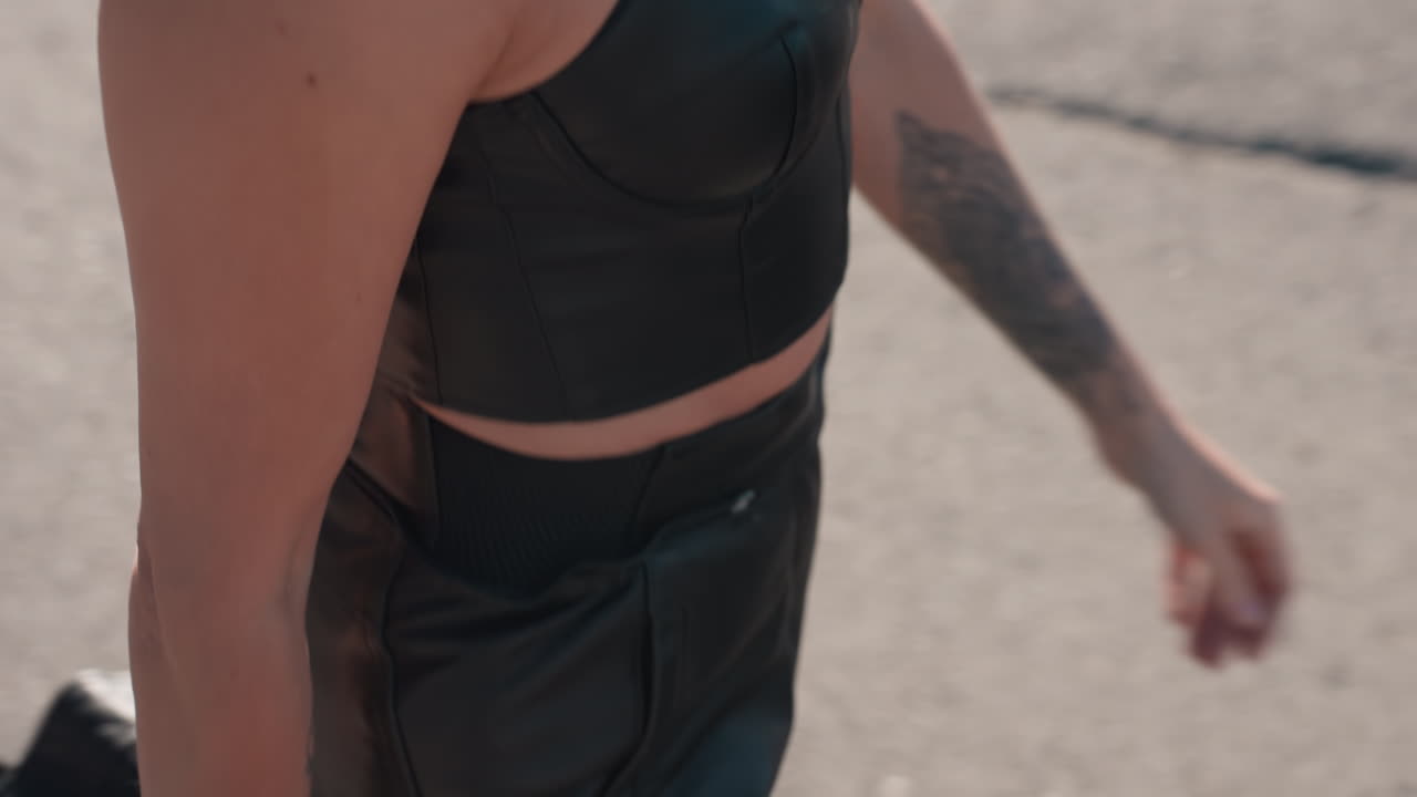 Close up of adult woman in black leather top and chain necklace holding sunglasses with tattooed hand while walking on tarred road, reflecting confidence, bold fashion and urban lifestyle
