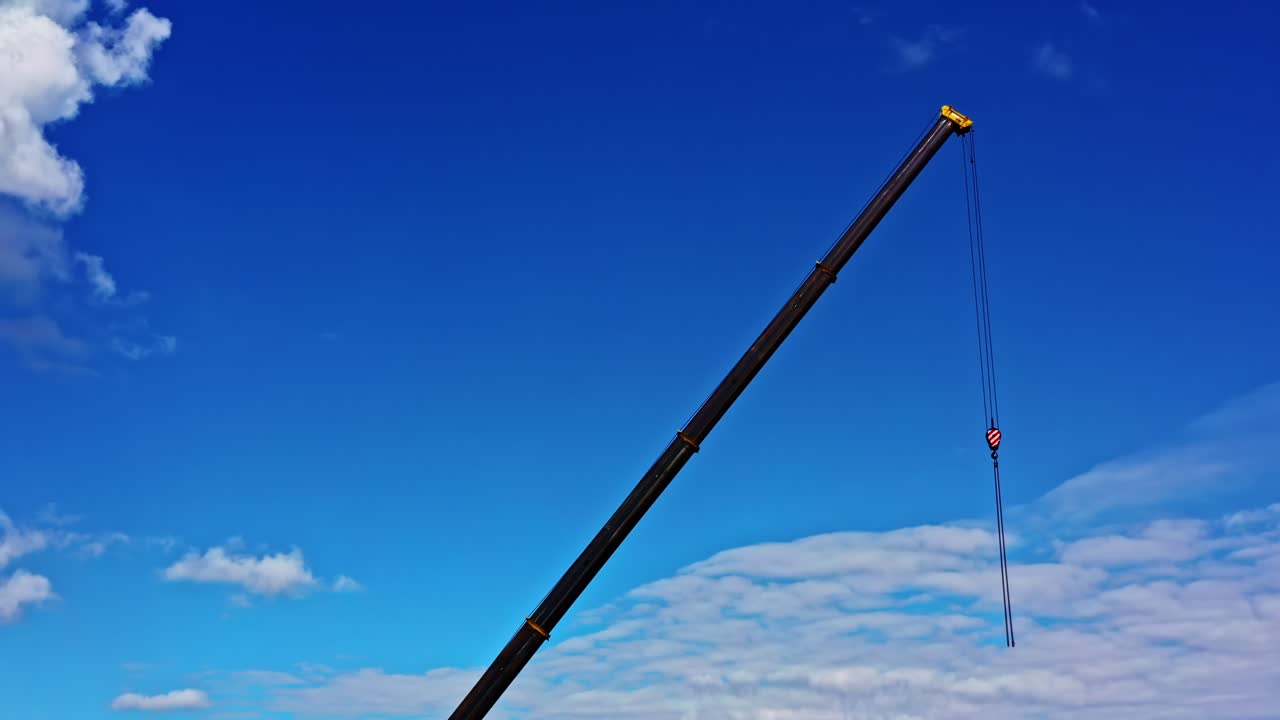 Telescopic crane boom with hook extended high against bright blue sky and clouds