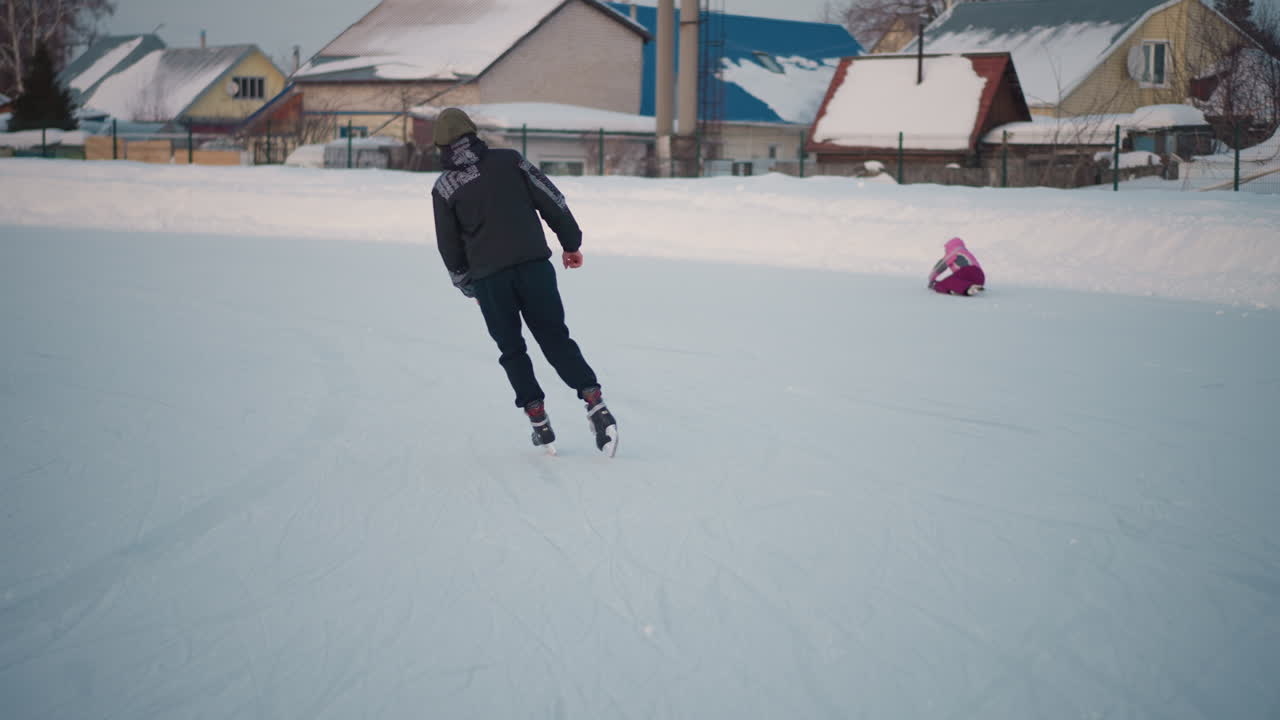 multiple people skating on frozen pond in winter jackets with one person in pink falling onto ice while others glide smoothly near snowy banks and houses in background under soft evening light