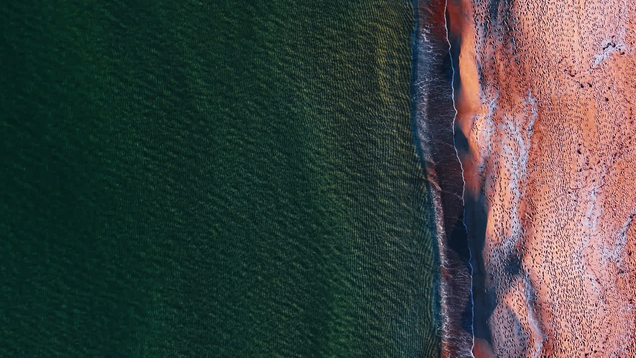 Topputsikt over havbølger som møter sandstrand med fotspor. Vertikalt flyperspektiv av grønt sjøvann og oransje sandstrand med tallrike fotspor.
