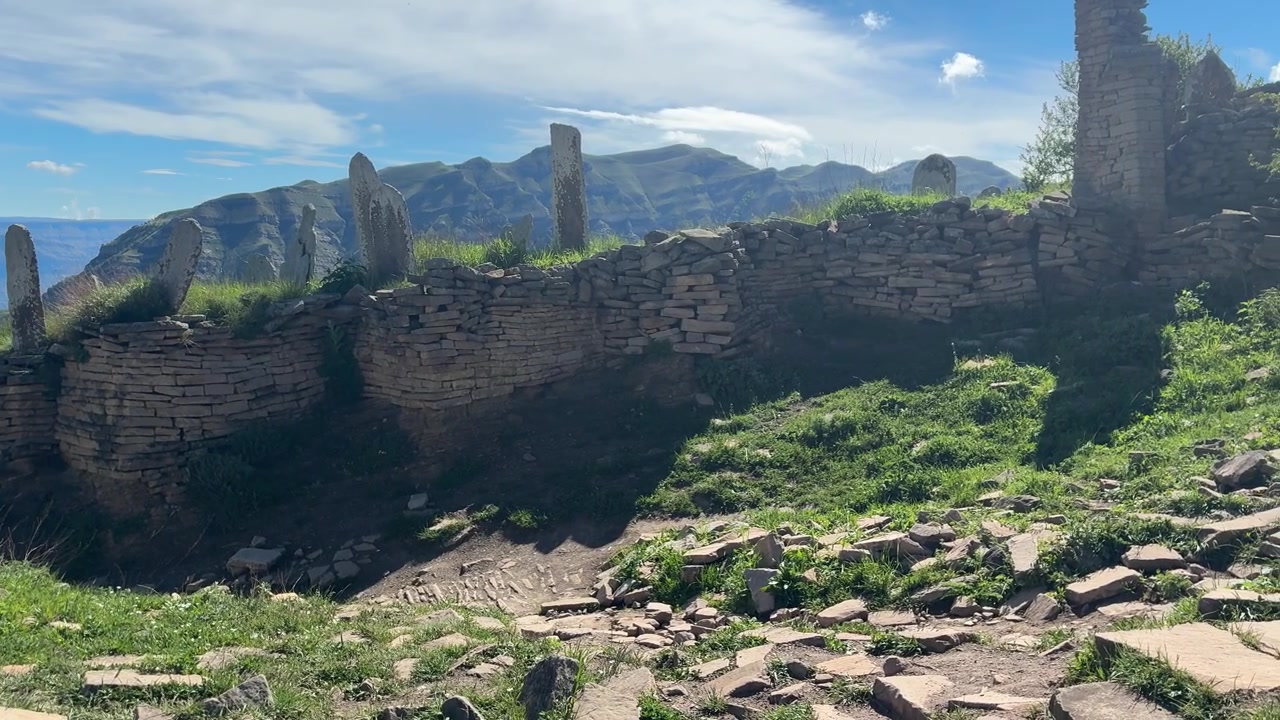 Uma tomada ampla captura as antigas ruínas de pedra de uma aldeia de montanha abandonada com relva verde e um céu azul nublado ao fundo.