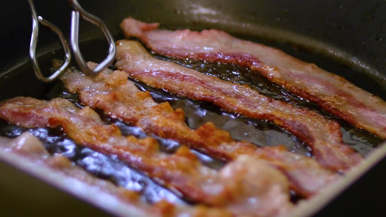 Bacon strips sizzling in frying are flipped by tongs, closeup