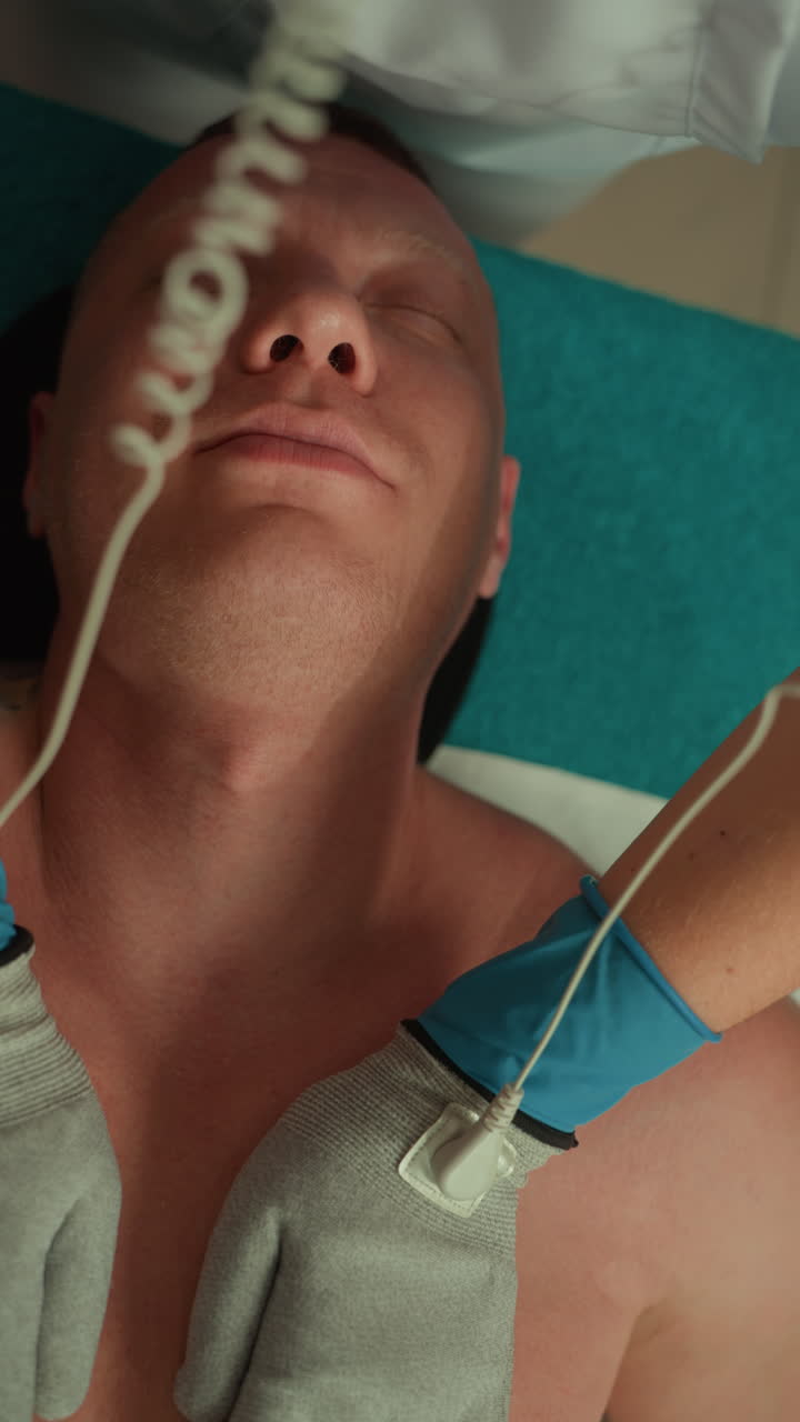 Kaukasischer Mann liegt mit geschlossenen Augen und erhält Mikrostrom-Gesichtsbehandlung, Kosmetikerin mit blauen Handschuhen gleitet mit Elektrodenpads über Kieferlinie und Hals, sanfte Impulse fördern Hautton und Beruhigung, türkisfarben