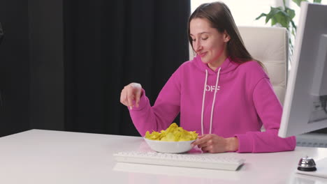 happy young woman sitting at desk and eating delicious potato chips