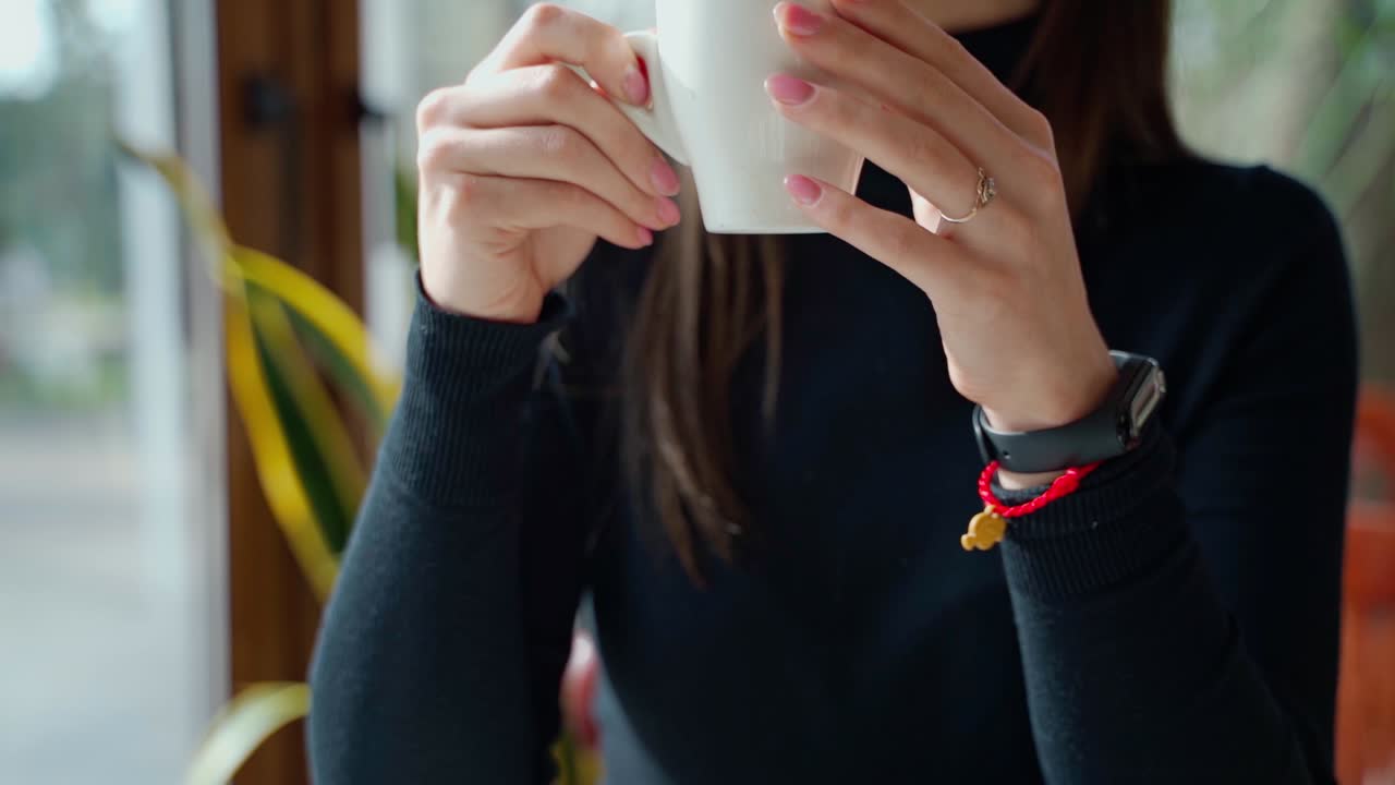Beautiful girl drinking coffee in a cafe. Young woman sitting at the table near the window and enjoys hot drink in the restaurant. Slow motion.