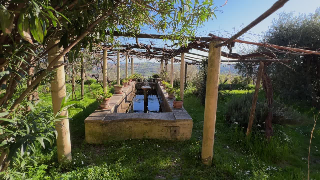 A long, ancient stone fountain stands under a rustic wooden pergola, surrounded by a green garden with views of the Umbrian countryside. Amelia, Italy