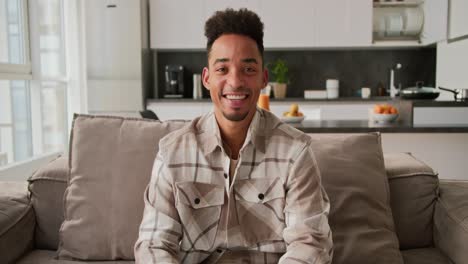 Portrait-of-a-happy-Black-skinned-brunette-man-with-stubble-in-a-beige-checkered-shirt-who-sits-on-a-modern-sofa-in-a-studio-apartment