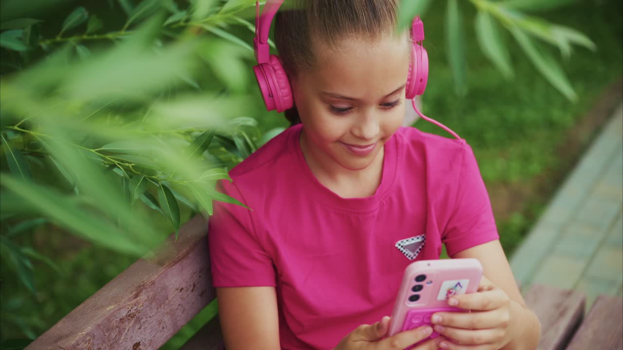 A Young Girl Enjoys Her Time Outdoors, Engrossed in an Entertainment Experience with Pink Headphones and a Smartphone Amidst Lush Greenery