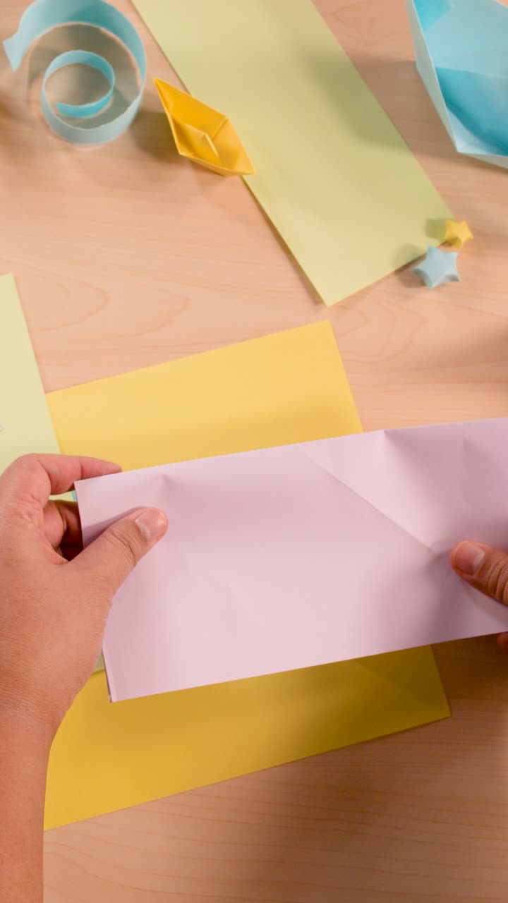 Mani piegano metodicamente carta pastello per origami, vista dall'alto, illuminazione soffusa, ambiente artigianale