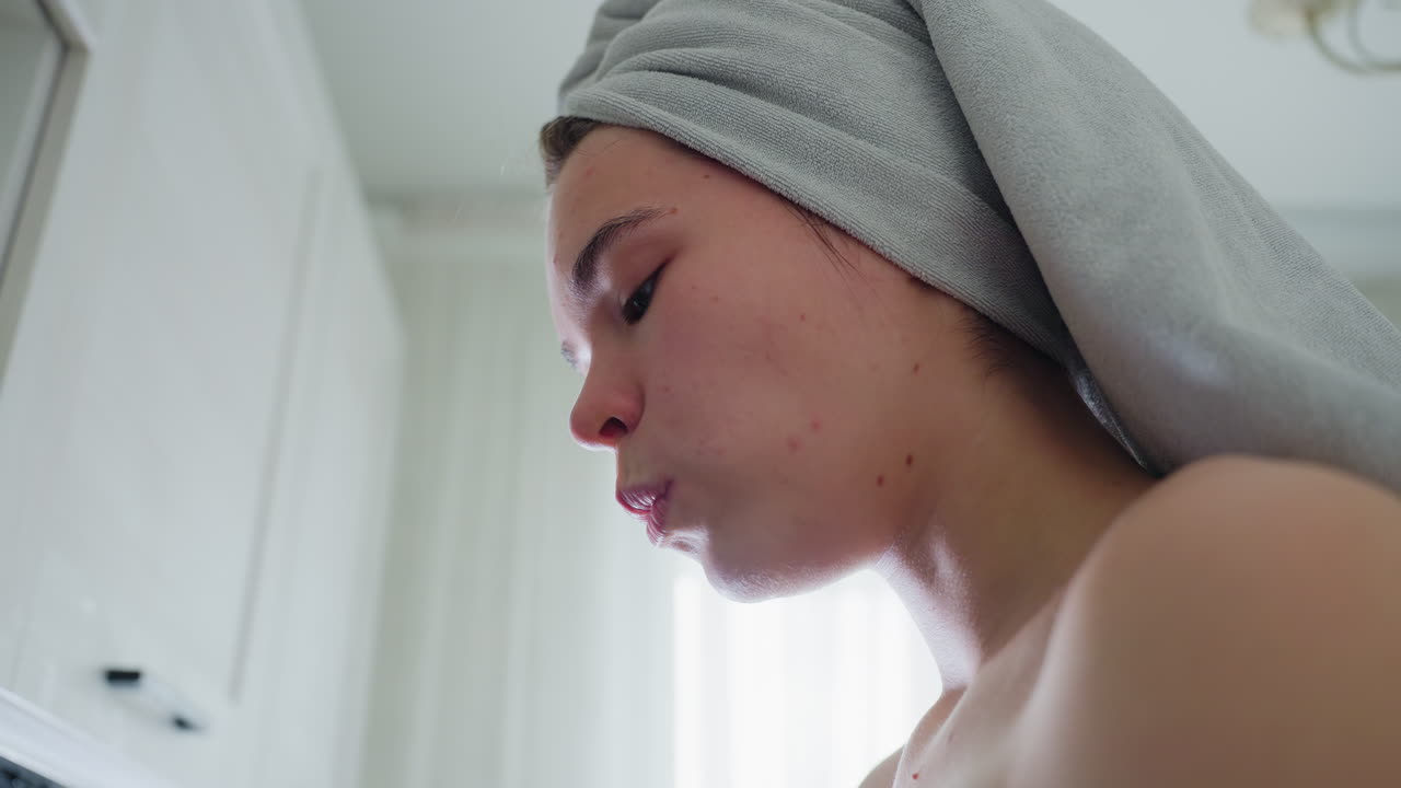 Close-up of woman with towel-wrapped head chewing cucumber slice near window, natural lighting illuminating relaxed expression and highlighting refreshing mood, wellness lifestyle