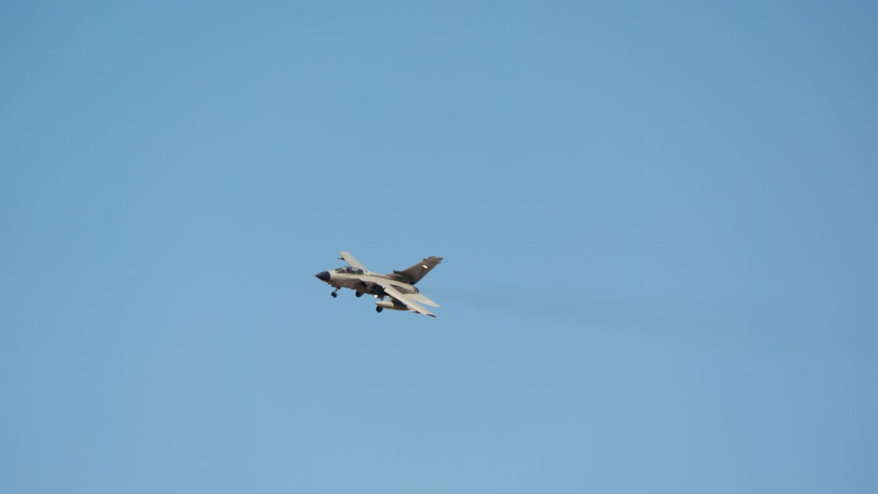 A military fighter jet with landing gear extended flies through a clear blue sky in slow motion as it approaches for landing