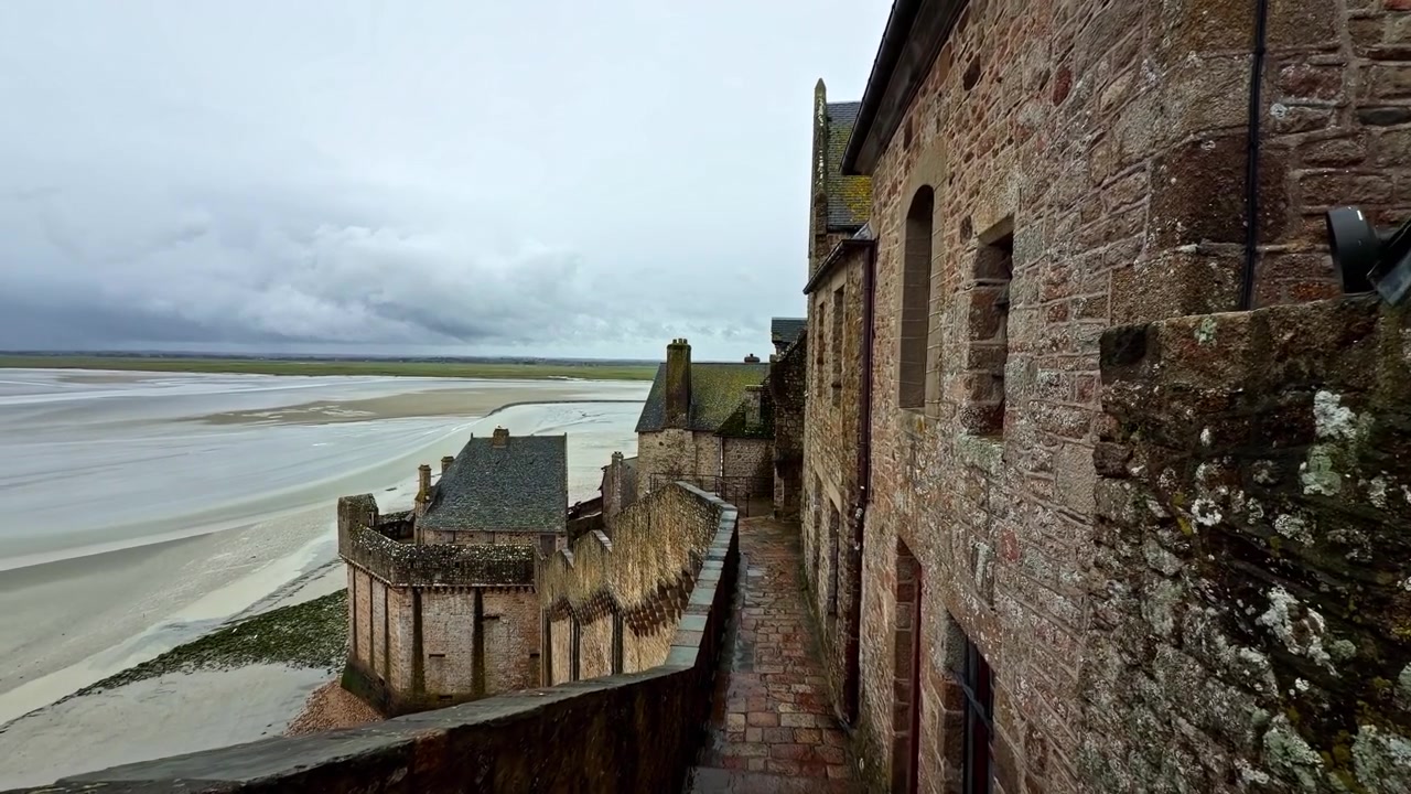 Những bức tường đá của Tu viện Mont-Saint-Michel trải dài về phía bãi triều rộng lớn dưới bầu trời nhiều mây. Hoàn hảo cho phim tài liệu lịch sử hoặc phóng sự du lịch