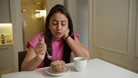 a sad brunette indian girl in a pink dress alone blows out a pink candle on her small cake and drinks coffee in a white mug while sitting at a white table in a modern apartment at home