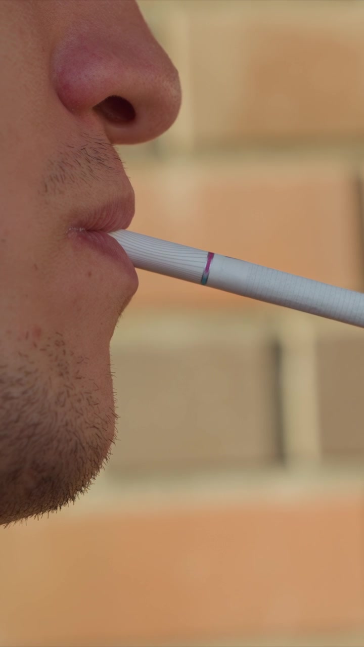 Primer plano de un hombre exhalando vapor de un cigarrillo electrónico. Las facciones del sujeto y un dispositivo de vapeo blanco con bandas de colores son visibles en todo el encuadre, con un fondo de pared de ladrillos difuminado.