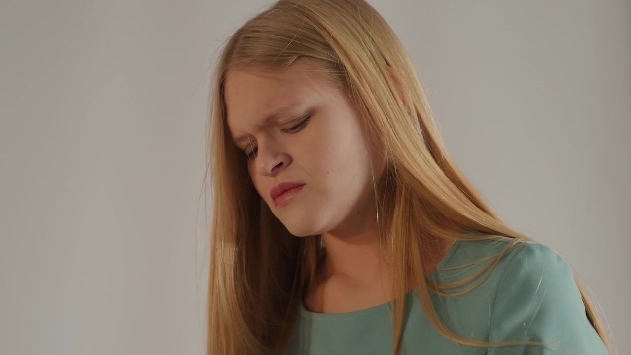 Young white woman portrait, pensive expression, actress rehearsing emotional scene in muted studio with turquoise blouse, long straight hair and downcast gaze, furrowed brows and soft lighting create
