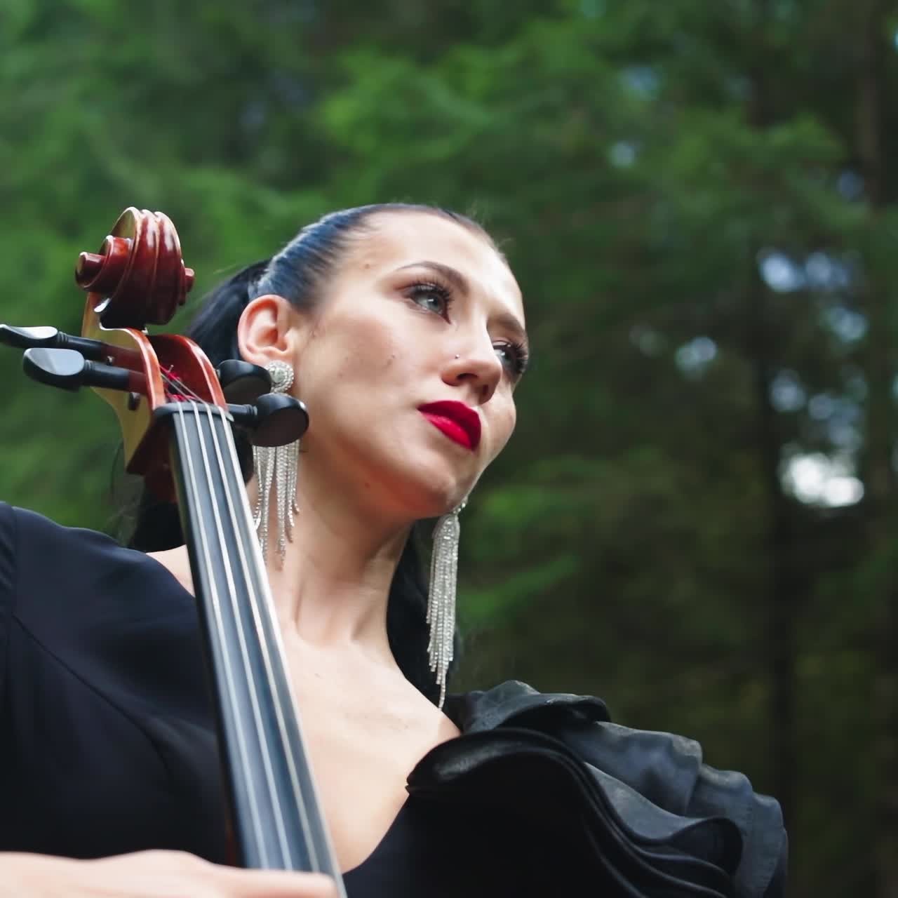 Attractive female musician on green nature background. Gorgeous woman in luxury earrings and dress playing the cello in the green forest.