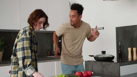 Happy-young-Black-brunette-man-in-a-cream-t-shirt-dances-with-his-young-adult-girlfriend-with-brown-hair-and-bob-hairstyle-while-they-are-preparing-breakfast-together-in-the-morning-in-a-modern-kitchen