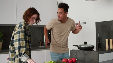 joven joven moreno feliz en una camiseta de crema baila con su joven novia adulta con cabello castaño y peinado bob mientras están preparando el desayuno juntos por la mañana en una cocina moderna