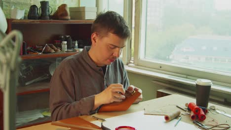 a young skilled tailor sews the element to a leather bag using a tailor's needle. in the interior of a leather workshop with a window