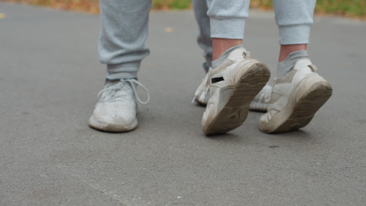 Leg view of two people wearing joggers and sneakers standing on pavement as one person raises feet stretching upward, indicating playful or affectionate moment outdoors with blurred background