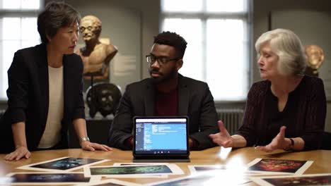 three individuals engaged in discussion around digital device and art prints in an academic setting, exploring ideas in history and culture