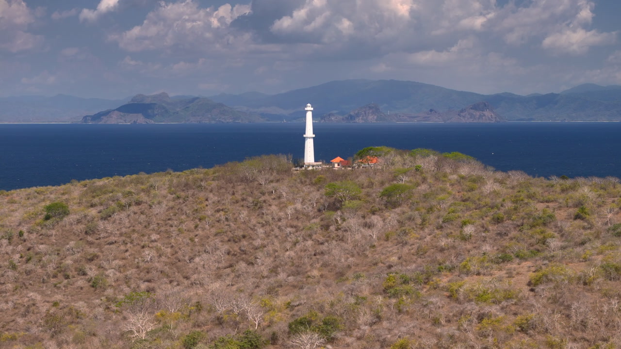 Aerial drone video of white lighthouse on dry coastal headland above deep blue sea with distant mountain island dramatic clouds and remote tropical landscape in bright daylight mountainous island