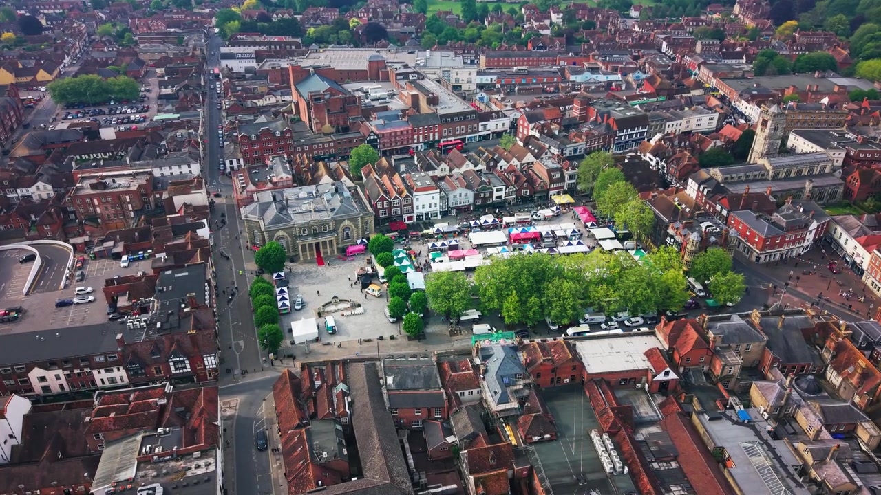 Droneopptak som ser ned over Salisbury Market Square med markedsboder, folkemengder, historiske bygninger, veier, trær og Salisbury-katedralen i det fjerne.