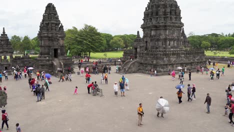 indonesia,prambanan temple on a cloudy day, camera tilts up slowly from the ground level and covering two temples, people are walking around the temple complex in slow motion.