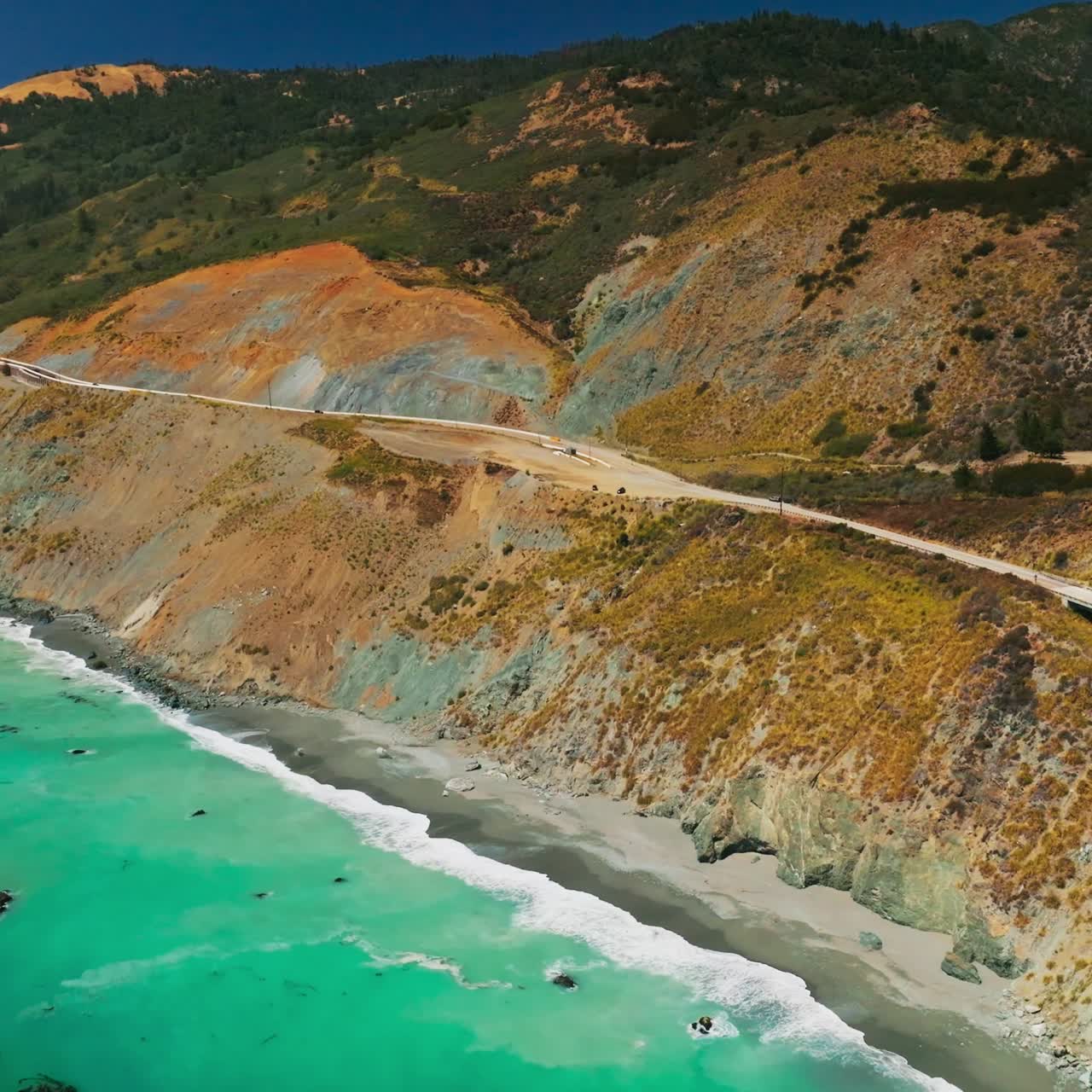 Amazing turquoise water of the Pacific with foamy waves arriving to the rocky shore. Mountains at the coastline with highway and bridge on scorching summer day. Top view