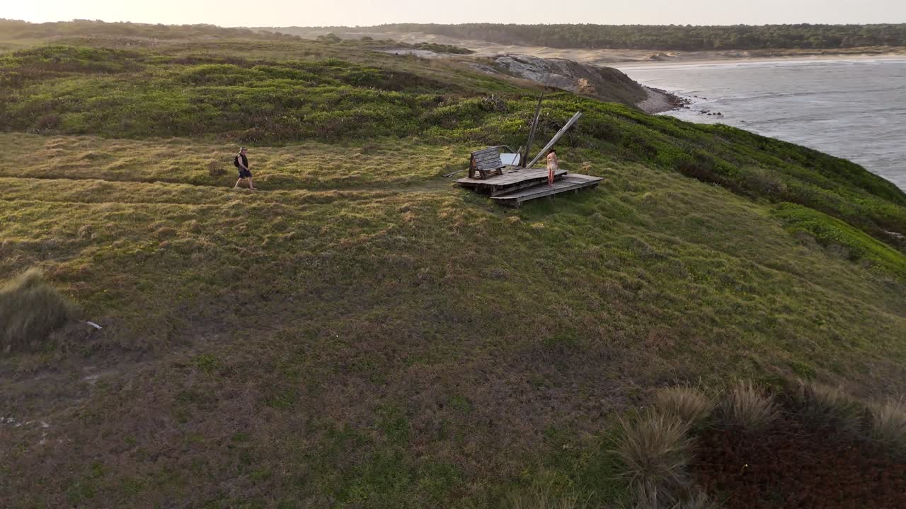 Vista aerea di un uomo che sale per raggiungere una donna in piedi su un punto panoramico che si affaccia sull'oceano al tramonto a Cerro Chato, Uruguay.