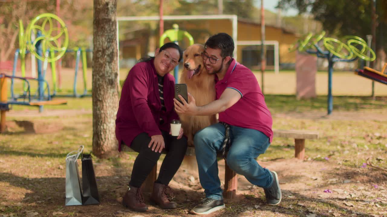 A man, woman, and their golden retriever dog take a selfie together on a park bench