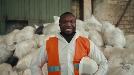 Close-up-portrait-of-a-happy-and-confident-man-with-Black-skin-in-a-white-protective-uniform-and-an-orange-vest-who-is-posing-near-a-pile-of-recycled-cellophane-and-plastic-at-a-waste-processing-and-sorting-plant