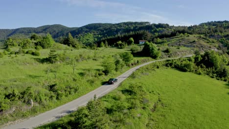 Toma-Aérea-De-Seguimiento-De-Un-Automóvil-Negro-En-La-Carretera-En-Las-Idílicas-Montañas-Verdes-De-Ródope-De-Bulgaria-Durante-El-Verano.