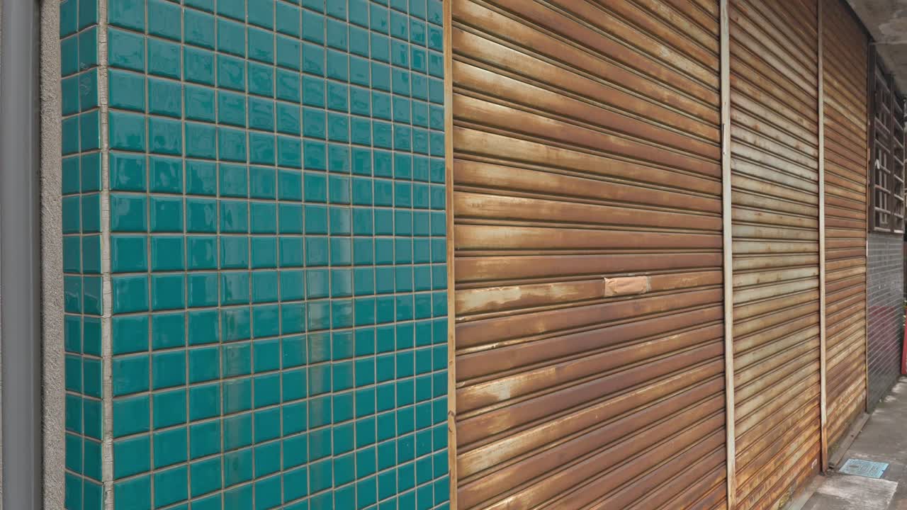 A textured facade featuring a vibrant turquoise tiled wall contrasting with weathered, rusting metal shutters, hinting at urban decay or disuse.