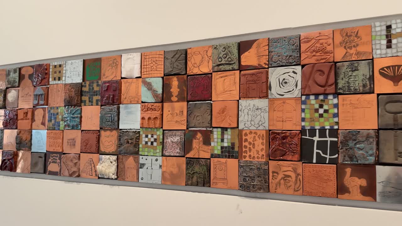 Una lenta panoramica su una lunga installazione artistica orizzontale con numerose piccole piastrelle di ceramica quadrate, dai design e texture diversi, esposte su una parete bianca