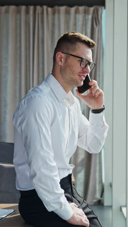 happy smiling man wearing official clothes and glasses in office. businessman speaking on the phone looking at window. vertical video