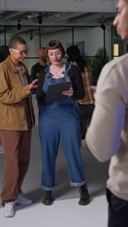 Vertical-Video-Of-Female-Film-Director-And-Assistant-On-Set-Shooting-Movie-Checking-Script-With-Crew-In-Background