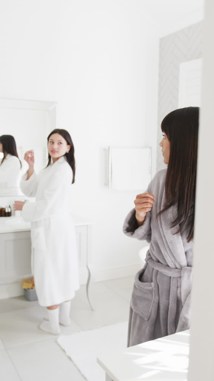 Vertical video, biracial mother and daughter in robes chat in sunny bathroom, slow motion