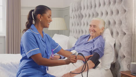 senior man at home in bed having blood pressure taken by female care worker in uniform