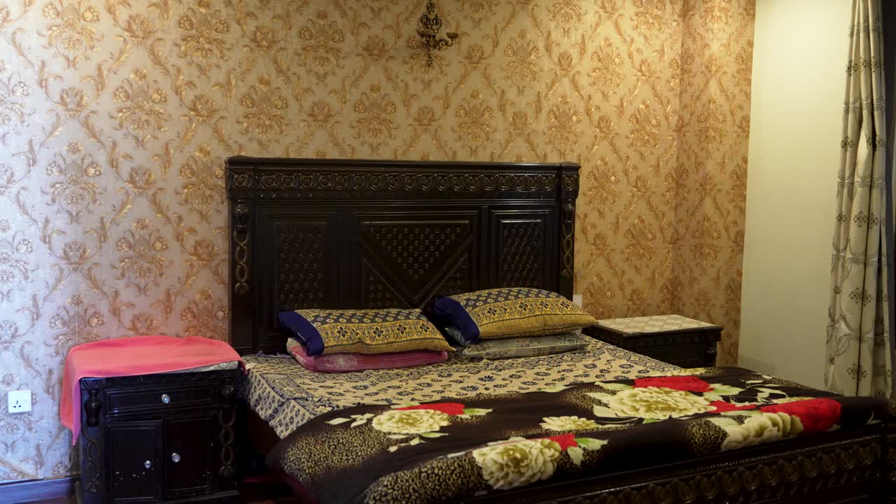 Tempat tidur kayu berukir dengan seprai bermotif bunga diletakkan di depan wallpaper bermotif di dalam kamar tidur.