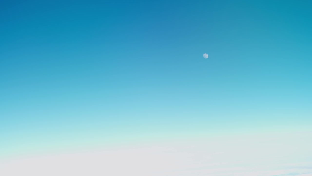 飛行中の雲の上に見える昼間の月と青い空、飛行機の乗客窓からの手持ち撮影