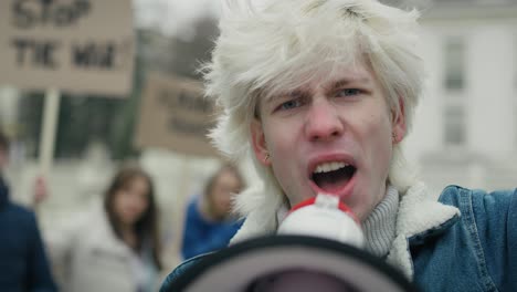 caucasian man looking at camera and screaming through the megaphone in front of manifesting  group of young people.