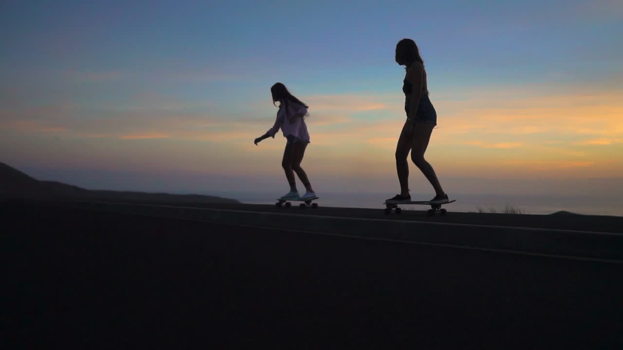 Two friends skate along a road at sunset, their movements captured in slow motion, against the backdrop of mountains and a breathtaking sky. They're wearing shorts