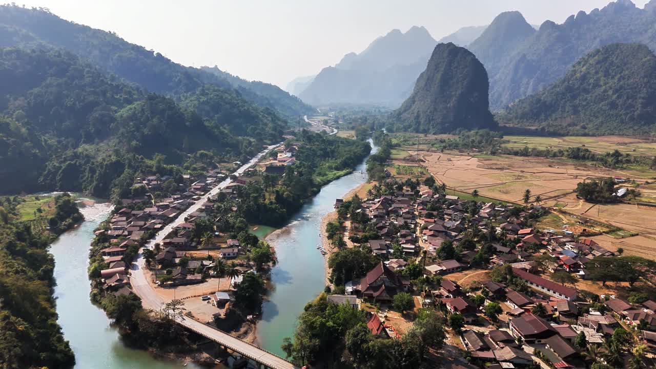 Aerial panorama of Pha Tang village in Vang Vieng, Laos, with a river flowing between traditional houses and dramatic karst mountains surrounding the peaceful rural landscape under daylight