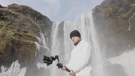 Videographer-Holding-Camera-on-Gimbal-Under-Waterfall-in-Landscape-of-Iceland-on-Sunny-Winter-Day